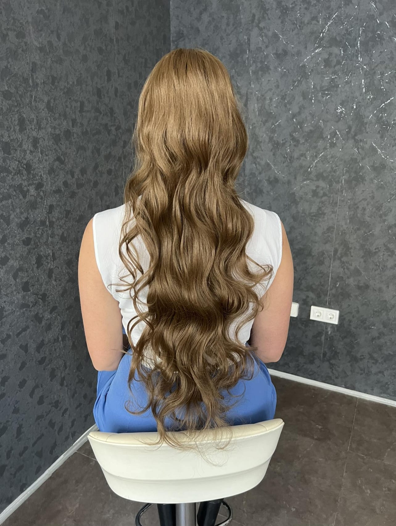 Rep za kosu - #10 smedji uvijeni 80cm - Umjetno vlakno 2 Duga talasasta kosa boje meda savršeno stilizovana na ženi koja sjedi na bijeloj stolici u modernom enterijeru Eynna hair Sarajevo